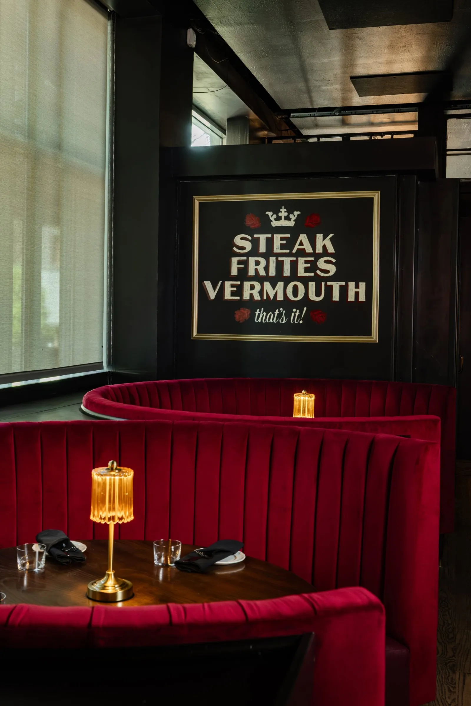 A red velvet booth with a brass lamp under the Steak Frites Vermouth sign